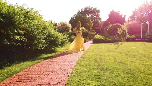 A blonde lady in a dress with is walking in the park. long train flutters and flies in the wind. Aerial video. Luxurious mysterious queen walks in the garden. Shooting from the back, without face