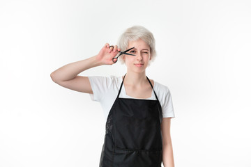 Pretty young hairdresser in white tee and black apron holding black scissors close to face ready for work standing isolated over white background. Wellness and beauty industry and professional career.