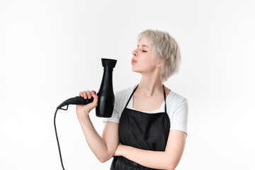 Young pretty hairdresser in white tee and black apron holding professional black hairdryer ready...