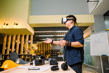 Young Asian man experiencing games with virtual reality goggles while standing in modern office. Headset VR device