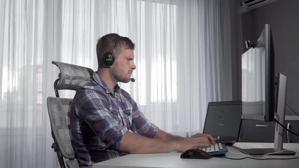 Handsome customer support operator typing on a computer. Charming young man working on the computer at call center, wearing headset with microphone. Consultant concept