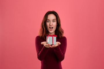 Excited pretty brunette girl holding present box with a ribbon with two hands standing isolated on a dark pink background and smiling at the camera. Celebrating special event.