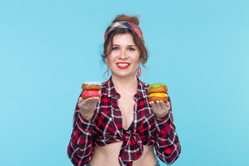 Positive young pin-up girl model holding in hands multicolored donuts posing on a blue background. Cooking concept for desserts and sweets.