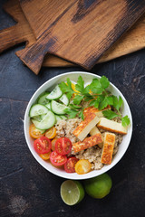 Bowl with quinoa, roasted cheese, cherry tomatoes, cucumber and parsley. Top view on a dark brown stone surface
