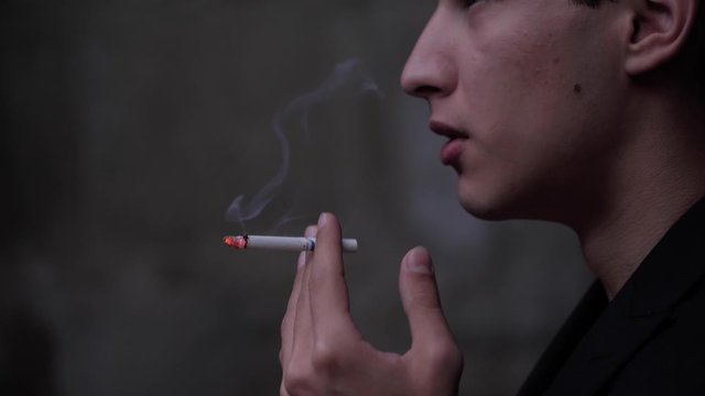 serious guy in black jacket smokes cigarette and blows white smoke cloud slow motion close view