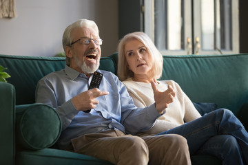 Annoyed elder woman looking at cheering senior husband, watching TV.