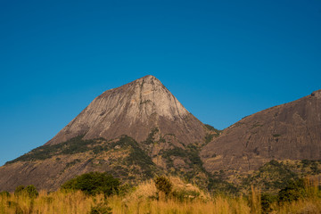 The rocky and conical shaped mountain rises steeply from the grassy plains under a clear blue sky in rural Mozambique, Africa