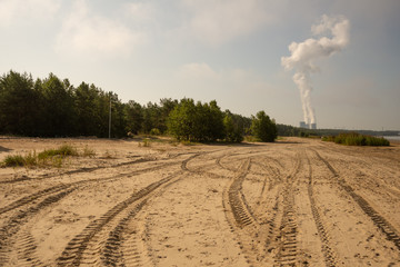 A beach with tire marks, steam from huge pipes creating clouds, a pine forest and the sea