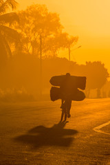 A backlit Mozambican cyclist with dramatic shadow, carrying a heavy load of coal to market along a tarmac road at sunrise. Nampula Town, Mozambique