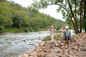 Mature female lifting trekking sticks and looking at her husband with camera