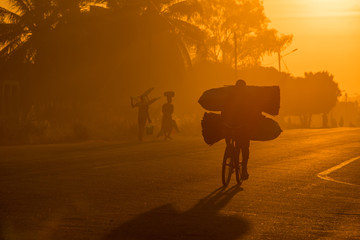 A Mozambican cyclist carrying large bags of coal to market at sunrise in the morning, along a tarmac road. Other commuters carrying items in the background. Nampula Town, Mozambique © Jennifer