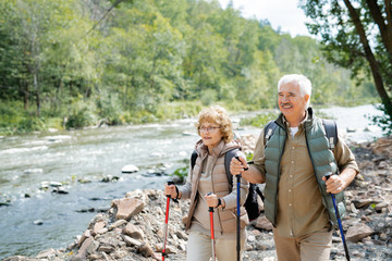 Naklejka premium Happy mature hikers with backpacks and trekking sticks walking along riverside