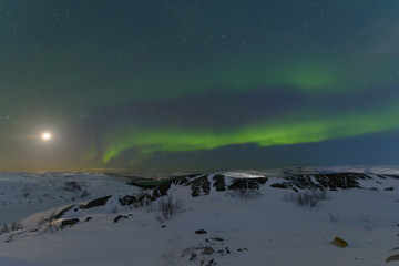 Fototapeta na wymiar Northern lights, aurora in the sky at night. Hills and rocks covered with snow. Moon in the sky.