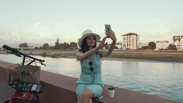 A young woman in a hat takes a selfie on the phone, a mountain river and a bicycle in the background. Girl on the promenade takes a selfie.