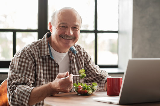 Smiling mature man having healthy breakfast sitting at the kitchen table with laptop computer at home and looking at camera