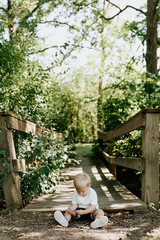 Naklejka na ściany i meble Cute Little Blond Haired Toddler Boy Kid Child Sitting and Laughing in Front of Wooden Bridge Over a Creek at the Outdoor Park in the Forest on a Sunny Pleasant and Happy Summer Day