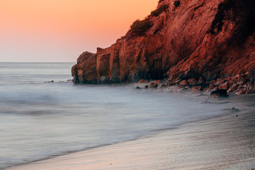 Long exposure photography of an ocean cliff as the waves come crashing into the beach. The water becomes silky smooth as the sunset darkens in color. California beach sunset, dramatic red and orange. - 288367805
