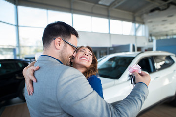 Handsome businessman husband holding keys and surprising his wife with a new car at vehicle dealership showroom. Happy people hugging and celebrating vehicle purchase.