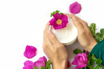 Young female hands are holding jar with white anti-ageing moisturizing cream with dog rose oil essential and vitamin E  on white background with bright pink dog roses, petals and green leaves