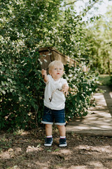 Cute Little Blond Haired Toddler Boy Kid Child Sitting and Laughing in Front of Wooden Bridge Over a Creek at the Outdoor Park in the Forest on a Sunny Pleasant and Happy Summer Day