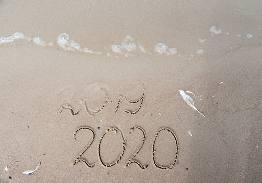 New Years 2020 replace 2019 concept. End of 2019 Happy New Year 2020, lettering on beach with wave and clear blue sea. Handwritten inscription 2019 and 2020 on beautiful sand beach.