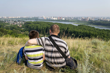 Father and son sit in a clearing on the mountain and look at the city.