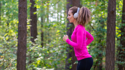 Woman jogging in a forest area and listening to the music via haedphones. Side view.
