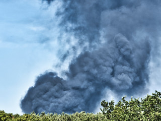 A Burning plastic recycling place causes a big pile of dark, black thick smoke on a sky blue summer day. Danger, sorrow. Terrorist attack or explosion.