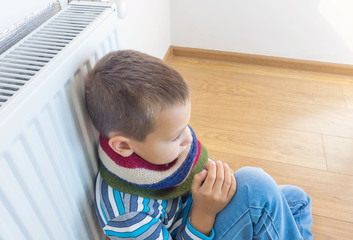 A young boy in a warm woolen sweater and scarf warms himself near a heating radiator at home. The...