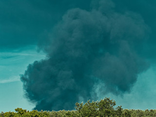 A Burning plastic recycling place causes a big pile of dark, black thick smoke on a sky blue summer day. Danger, sorrow. Terrorist attack or explosion.