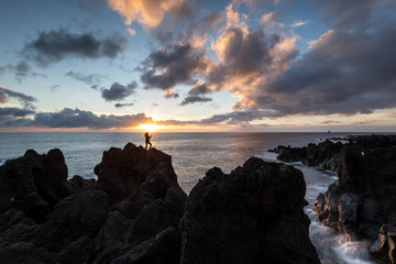 Young woman enjoying a beautiful sunset at the most western point of Europe Fajã Grande from the rocks of the black lava coast