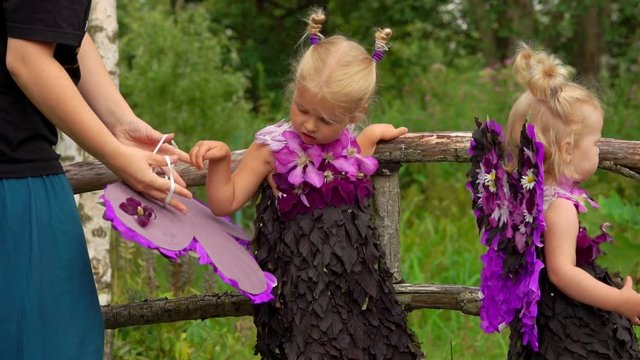 Mom helps the girl to put on the wings of a butterfly. Children in fairy costumes play in the garden.