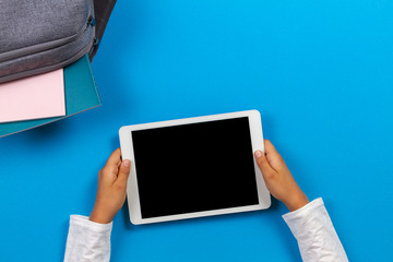 Top view to kid hands with tablet computer, backpack with books and notebook on light blue background