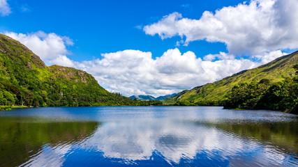 Beautiful shot of a tranquil lake and mountain with puffy clouds reflected in water. Scenic green...