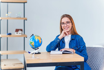 Student girl looks for window and dreams about vacations while studying at white living room. Studying geography with globe. Cute schoolgirl in denim clothes makes hometask.