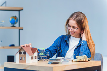 Young pretty girl counts her savings money and looks to a model of the house.