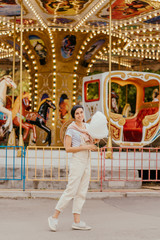 Young woman  with  cotton candy in front of the carrousel with night illumination at the amusement park.