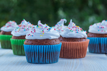 Delicious muffins with white cream and colored sprinkles on a wooden table on a background of green foliage.