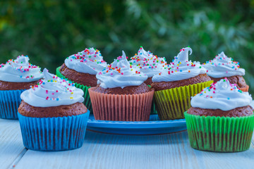 Delicious muffins with white cream and colored sprinkles on a wooden table on a background of green foliage.