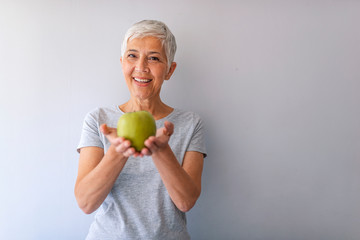 Close up of a cheerful elderly woman eating an apple while smiling. Portrait of senior woman with green apple, indoors, with copyspace. Portrait of happy mature woman holding granny smith apple