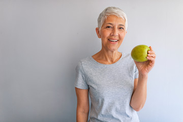 Older woman with healthy food indoors. Smiling woman eating green apple. Portrait of a beautiful...