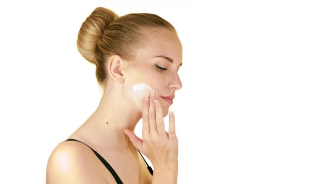 Close-up static slow-motion shot of a beautiful middle aged blonde lady rubbing moisturizer cream on har face gently with her fingers while looking sensually at the camera with a white background