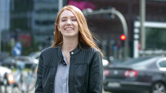 Close Up: Cheerful young girl with red hair is smiling to the camera. In the background blurred street, traffic lights and cars.