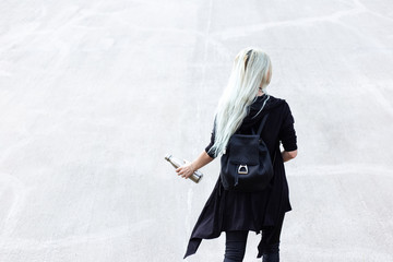 Back view of young blonde girl, dressed in black with backpack, holding steel thermo bottle in hand. On background of textured light grey wall.