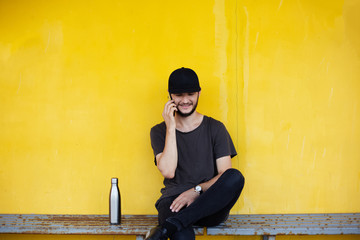 Smiling guy is sitting on yellow background near steel thermo bottle and talking on smartphone.