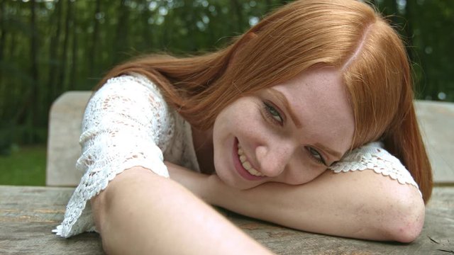 Beautiful happy red haired girl sitting on a park bench and smiling in the camera. Slow Motion Close Up Footage.