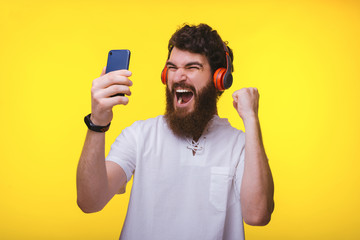 Excited bearded guy, with headphones, holding a mobile phone and celebrating the won, over yellow backgrdound