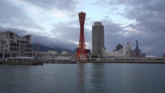 Sunset view of Kobe, Japan waterfront with buildings and city skyline across the bay. Dramatic sky and clouds form a peaceful evening scene over the city. Modern Japanese architecture.