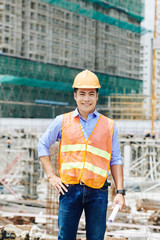 Fototapeta na wymiar Portrait of positive Vietnamese head engineer in bright vest standing with blueprint scroll at construction site