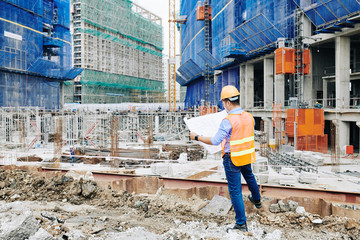 Rear view of head engineer in orange vest examining building blueprint when standing at construction site